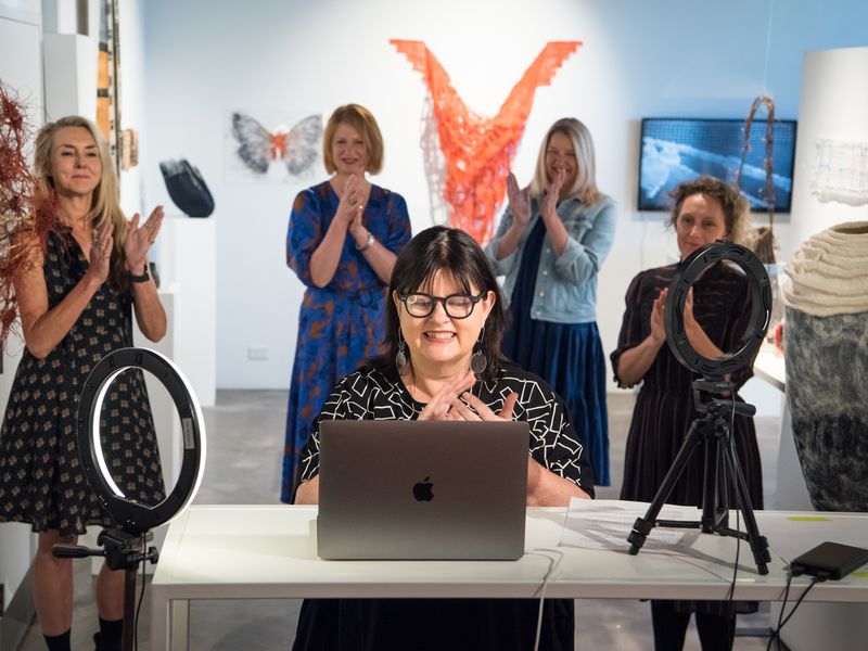 Photo of a woman with lights speaking into a computer for a zoom opening with four women standing  behind in a gallery space.